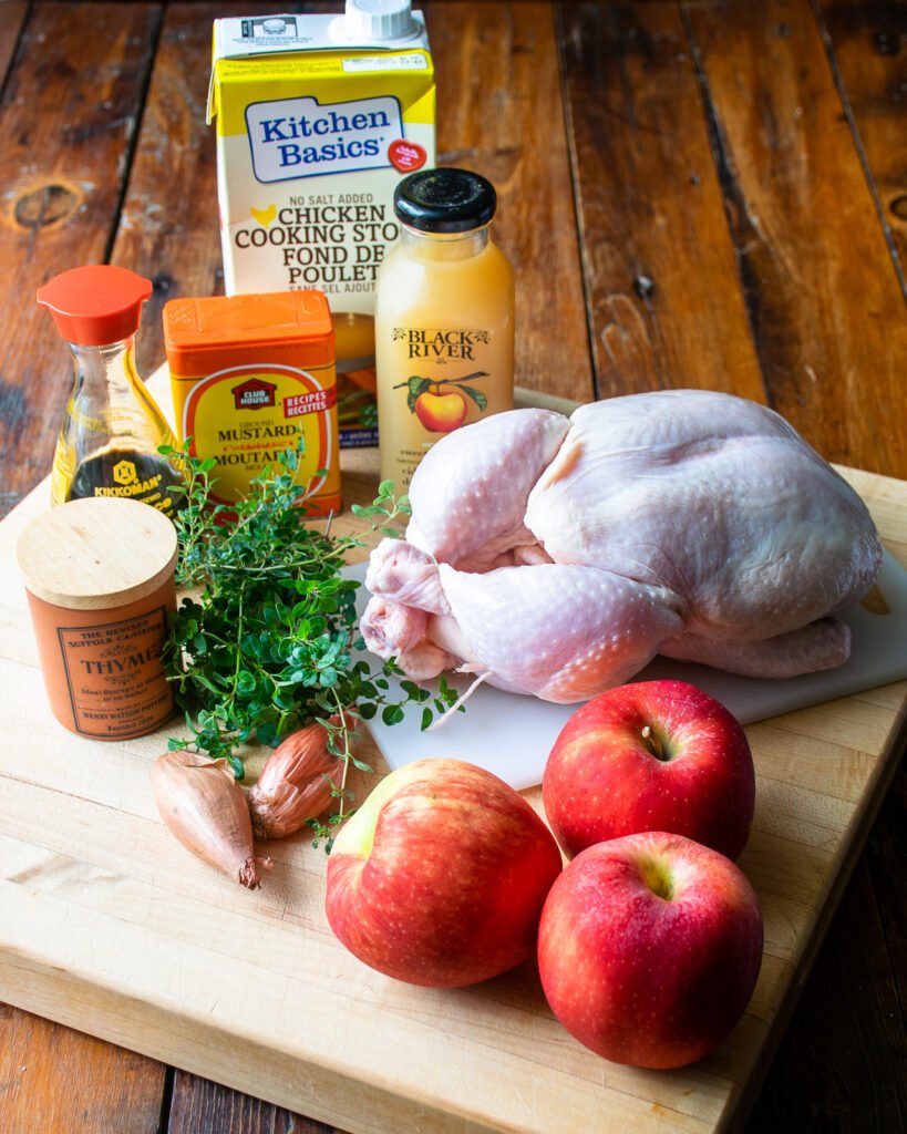 Ingredients on a cutting board - a whole chicken, apples, shallots, thyme, dry mustard powder, apple cider, soy sauce and chicken stock.
