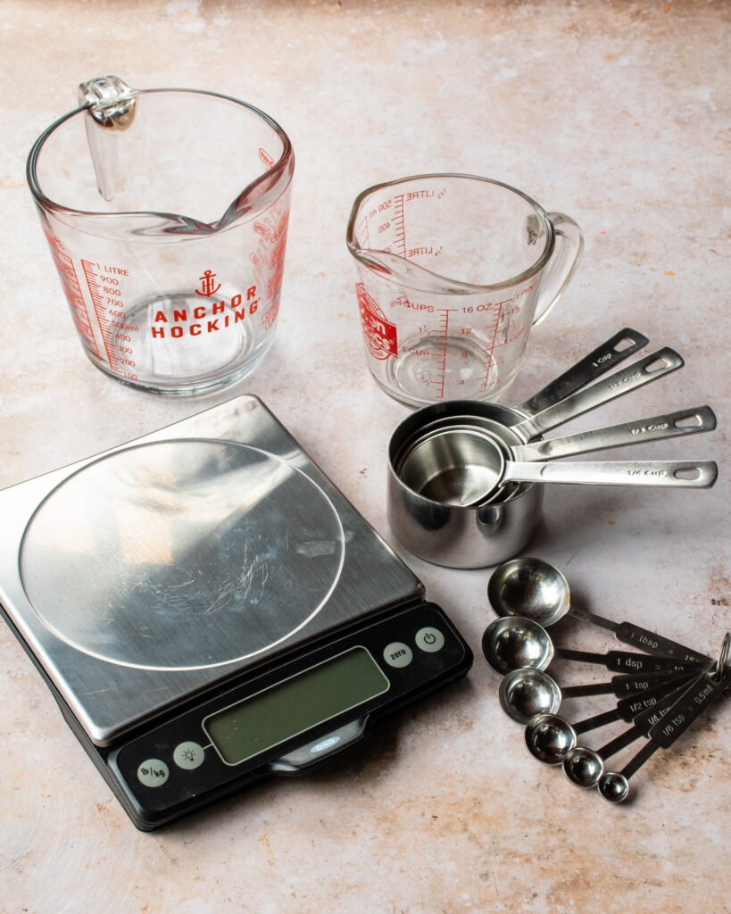 A scale, two glass measures, dry ingredient cup measures and a set of measuring spoons on a countertop.