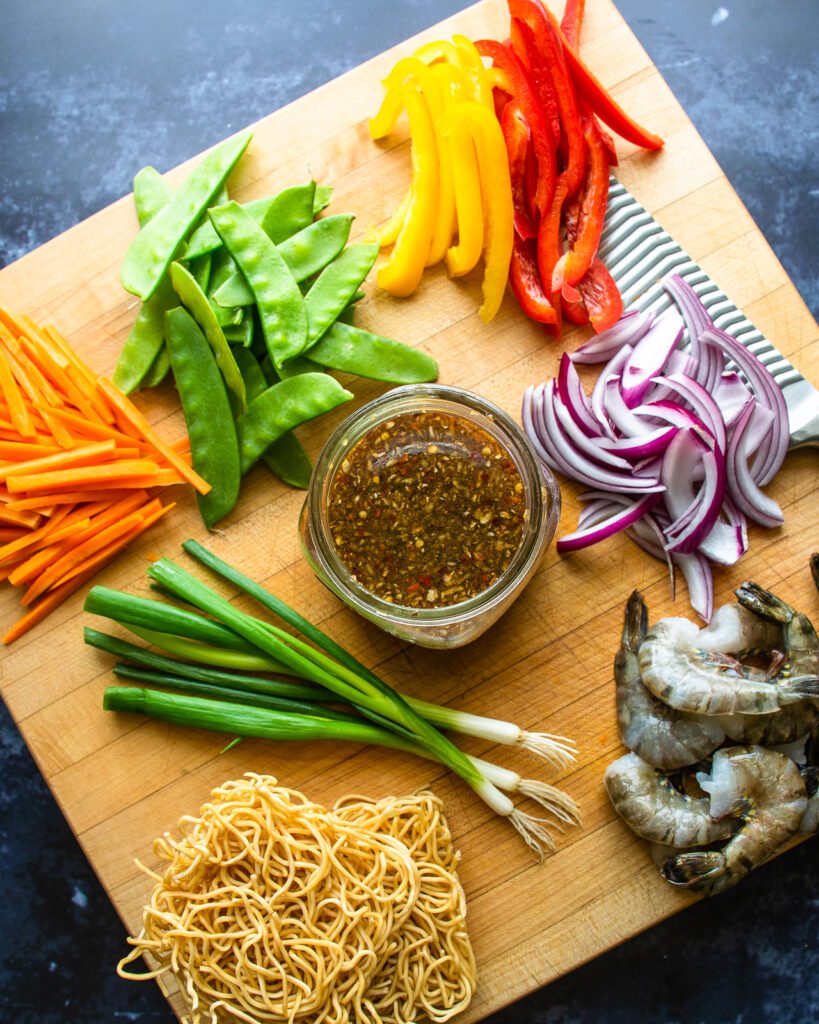 Chopped vegetables on a cutting board along with dried noodles, raw shrimp and a jar of stir fry sauce.