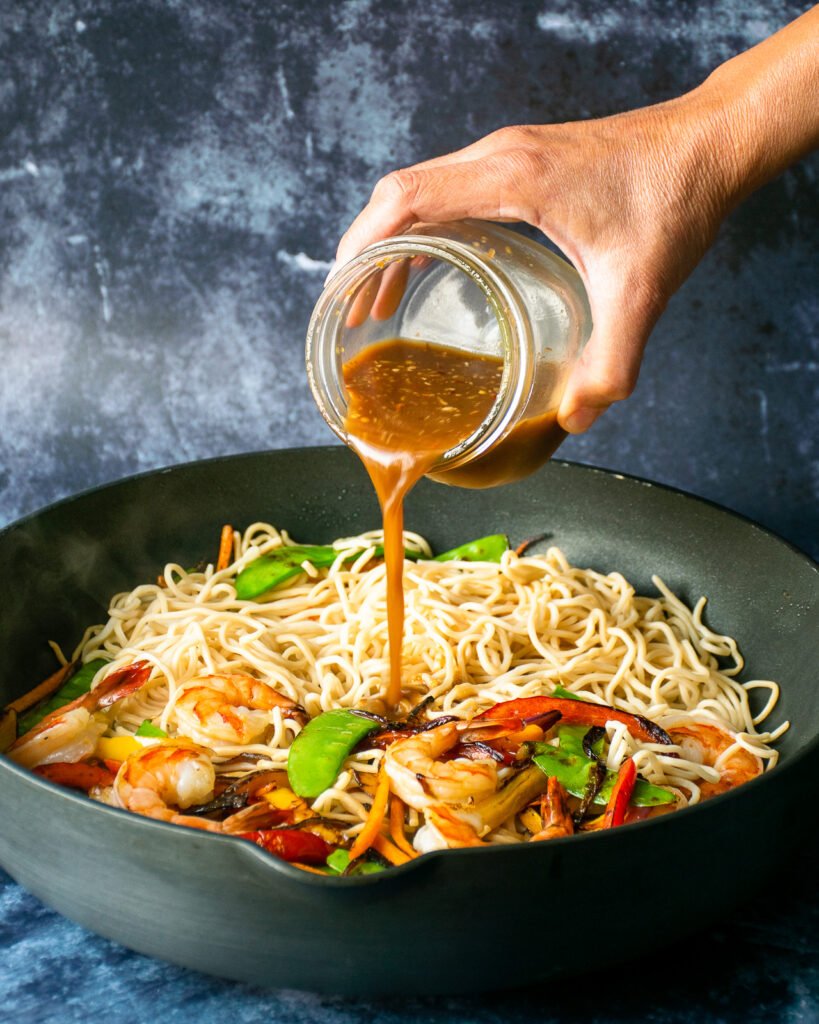 A hand pouring stir fry sauce over vegetables, shrimp and noodles in a large skillet.