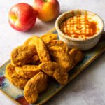 Apple fries on a plate with caramel cream dip and two apples next to it.