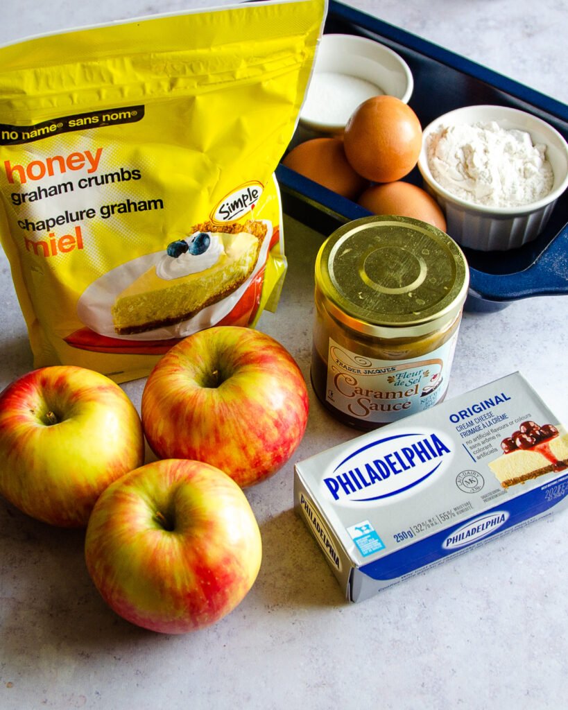 Ingredients on a counter - apples, cream cheese, caramel sauce, eggs, graham cracker crumbs, sugar and flour.