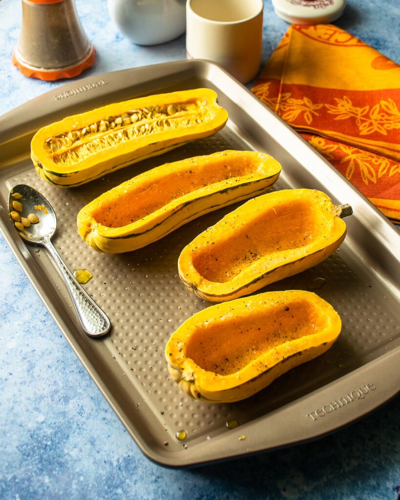 Four halves of delicata squash on a baking sheet - three with seeds scooped out and one with seeds still in.