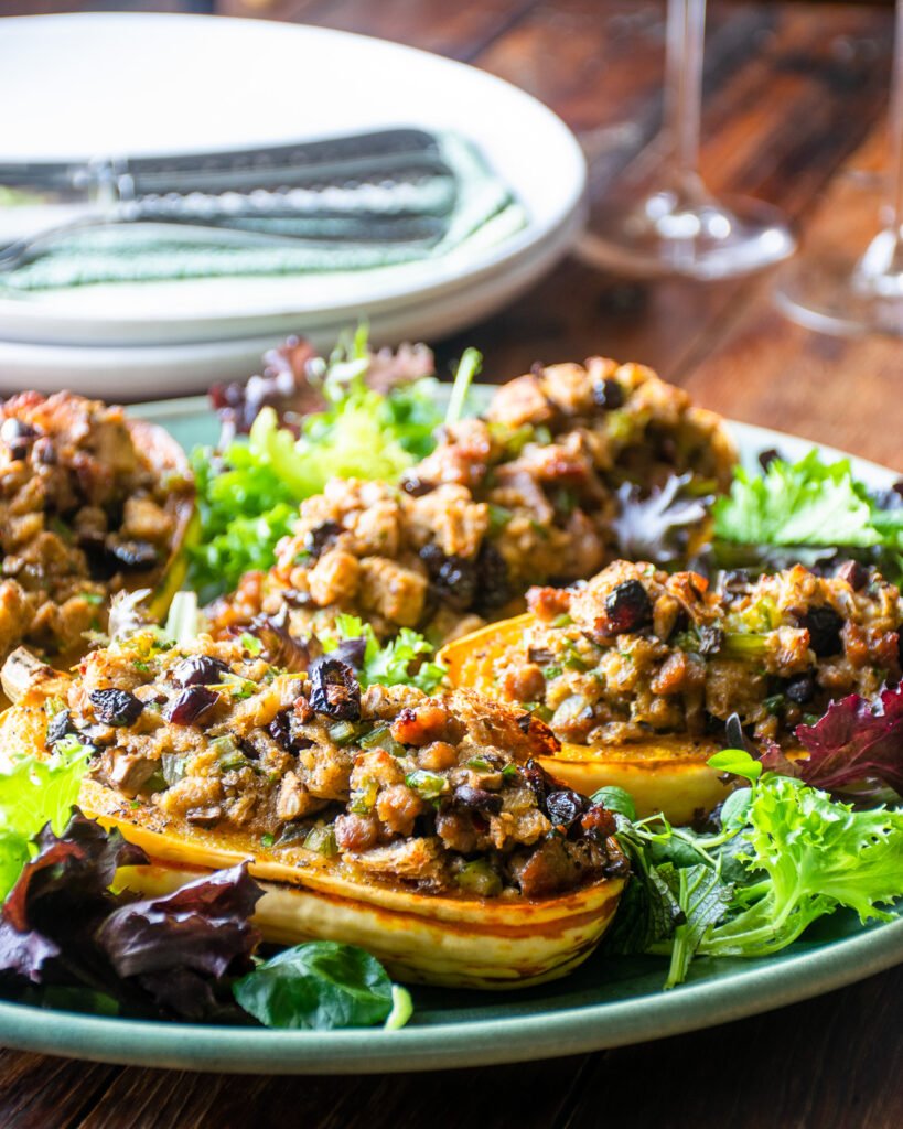 Stuffed delicata squash on a platter with greens, with plates and wine glasses in the background.