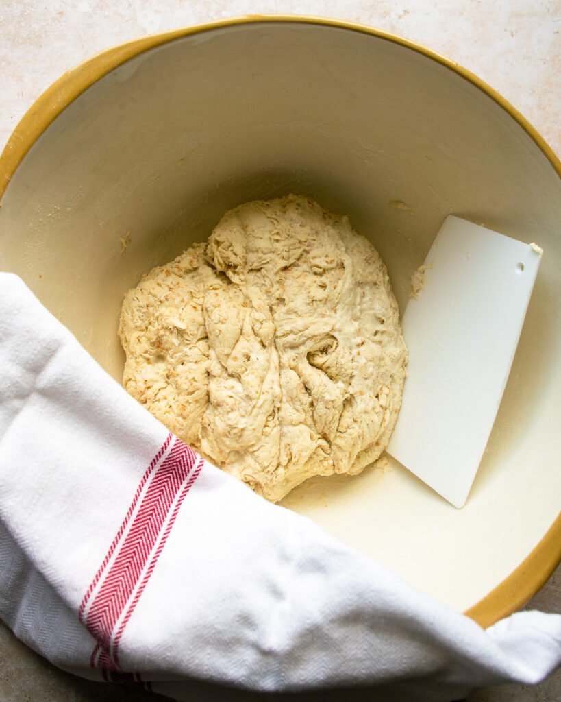 Sourdough bread dough in a large bowl with a red striped towel partially covering it and a plastic board scraper.