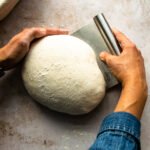 Two hands shaping sourdough bread with a bench scraper.