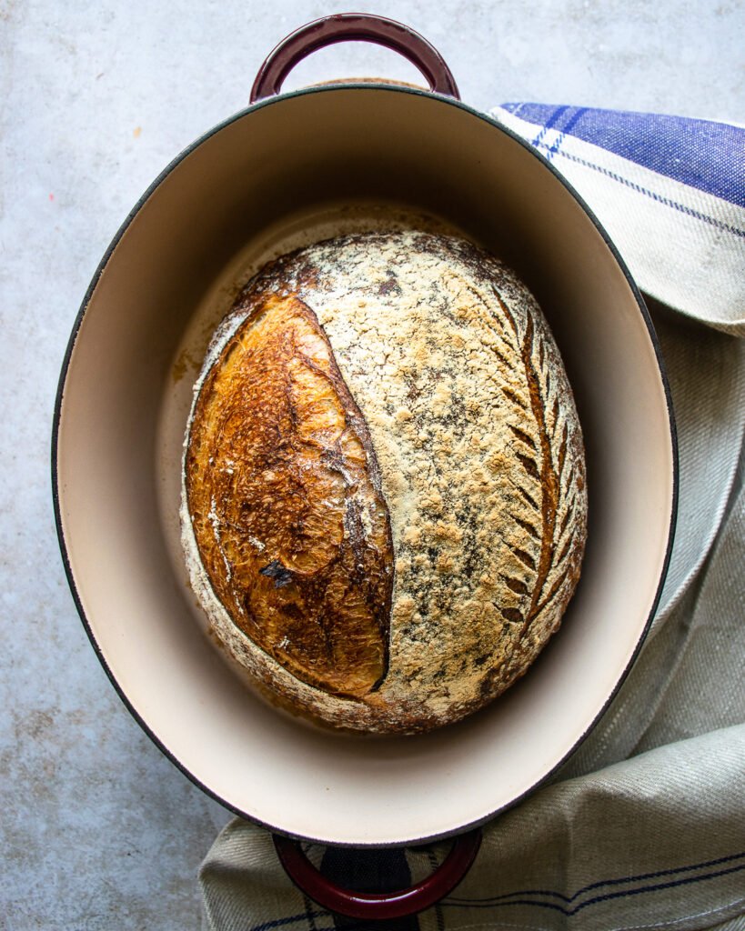 A loaf of baked sourdough bread in a cast iron pot with a towel next to it.