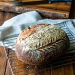 A loaf of sourdough bread on a cooling rack with a kitchen towel, a bread knife and cutting board in the background.