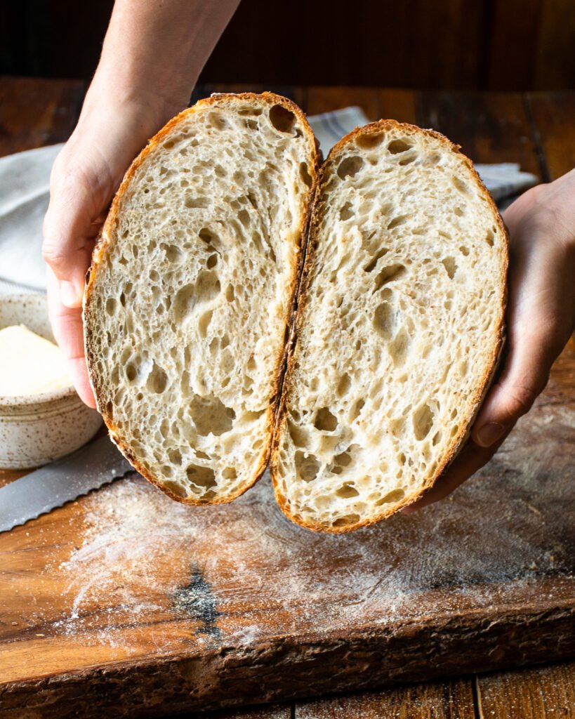 A loaf of sourdough, cut in half, held in two hands over a cutting board.