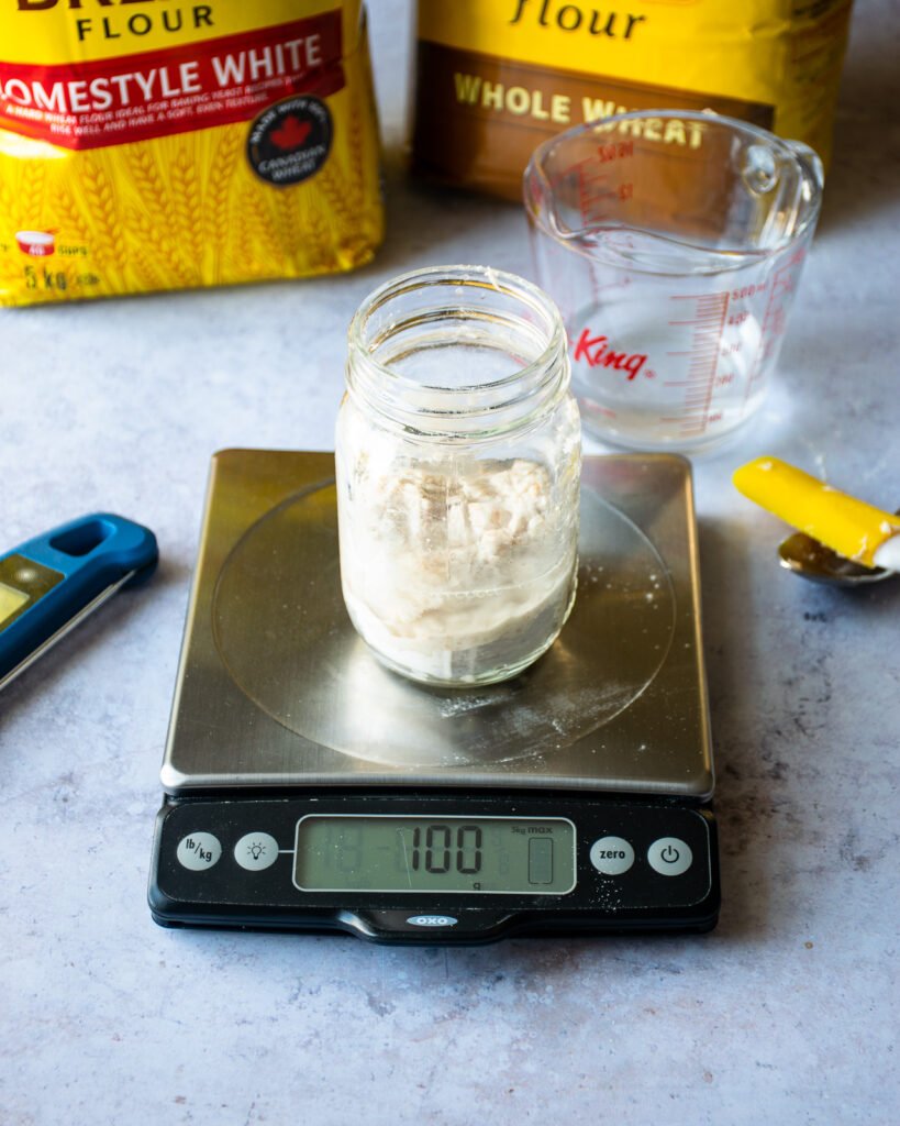 A mason jar on a scale with flour and water inside, next to two bags of flour, a thermometer and a spatula.