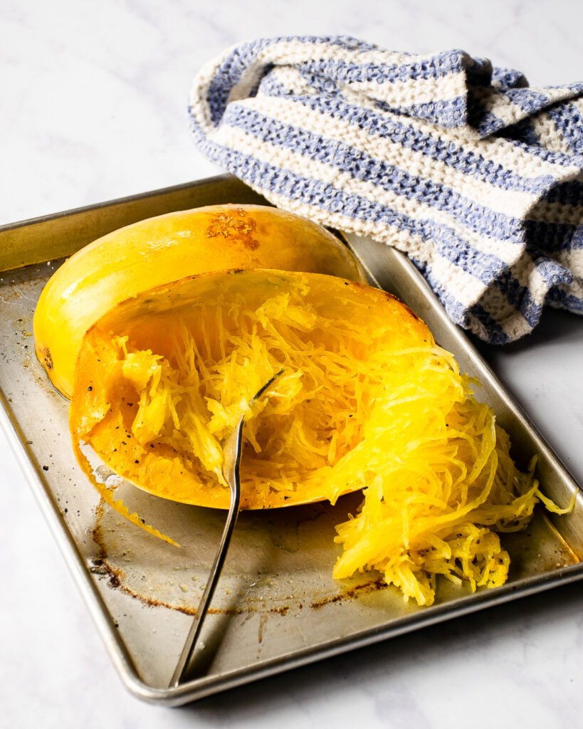 A fork shredding spaghetti squash on a baking sheet with a blue and white striped towel.