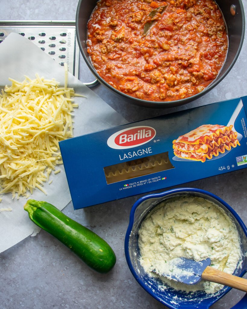 The components of lasagna on a countertop - bolognese sauce, grated cheese, pasta, ricotta and zucchini.