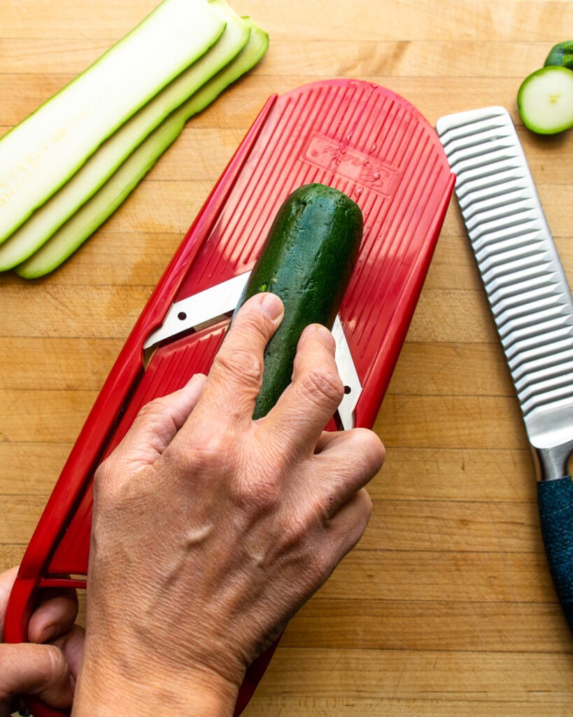 A hand slicing zucchini into long strips on a mandolin slicer.