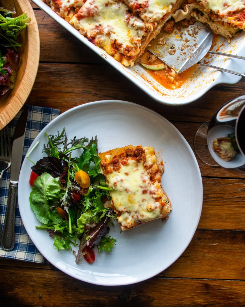 Overhead shot of a white plate with a piece of lasagna with zucchini and a side salad.