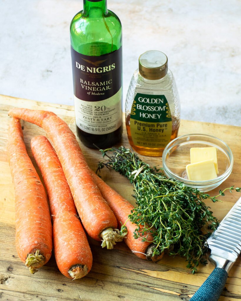 Ingredients on a cutting board - carrots, thyme, balsamic vinegar, honey, butter and a knife.