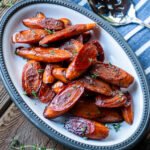 Balsamic glazed carrots on a white oval plate on a wooden table with a blue striped napkin and serving spoon.