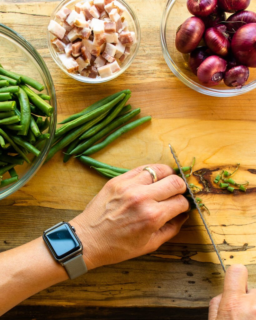 Hands cutting the ends off green beans with a bowl of onions and a bowl of chopped bacon near by.