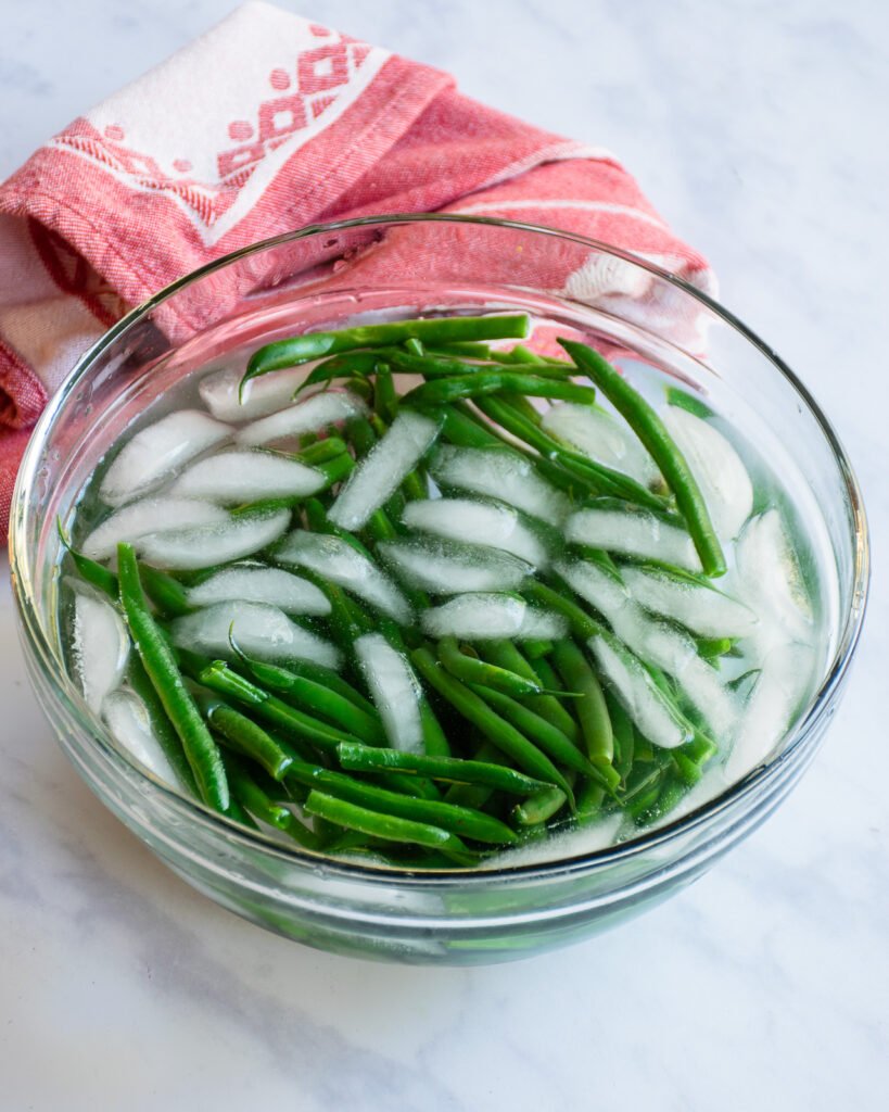 Green beans in an ice bath with a red kitchen towel behind it on a marble counter.