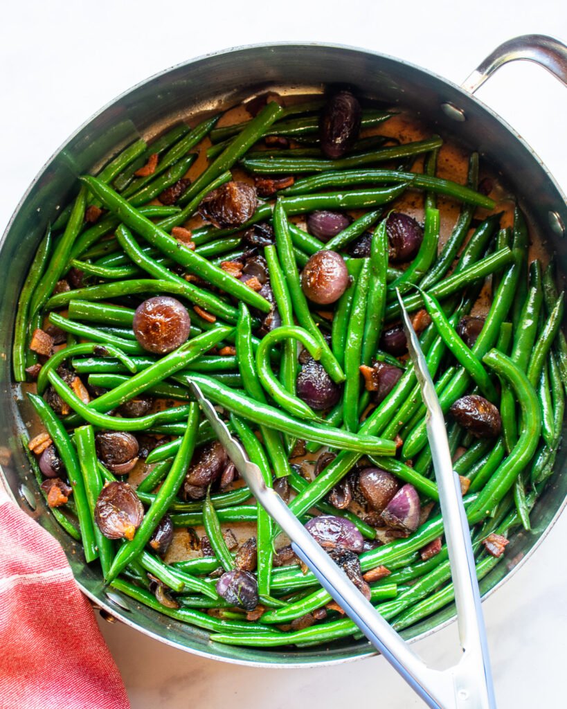 Green beans with onions and bacon in a straight-sided saute pan with tongs and a red kitchen towel.