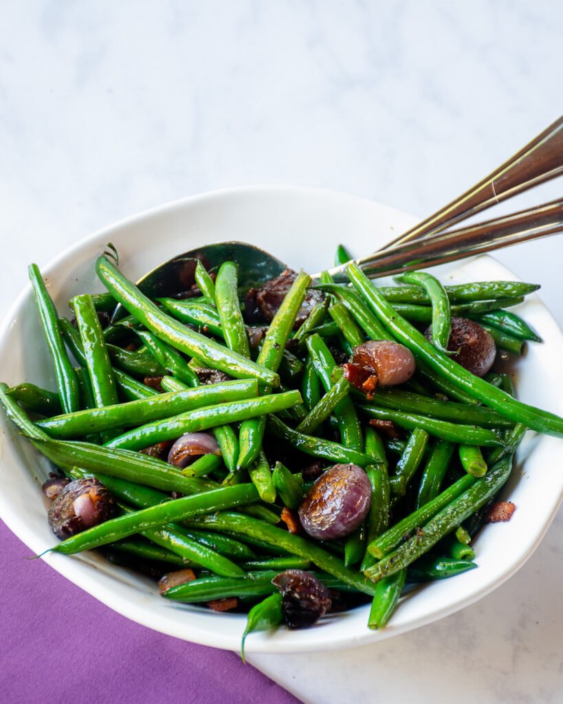 Green beans with onions and bacon in a white bowl with serving utensils and a purple napkin.