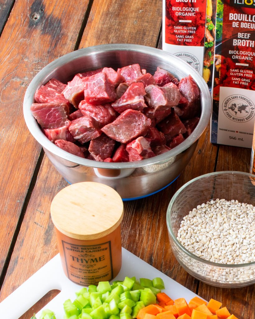 A bowl of stew beef on a wooden table with herbs, vegetables, beef stock and a bowl of raw barley.