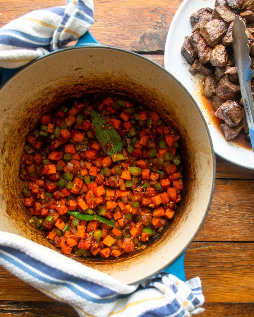 Vegetables cooking in a cast iron Dutch oven with a plate of browned beef next to it.