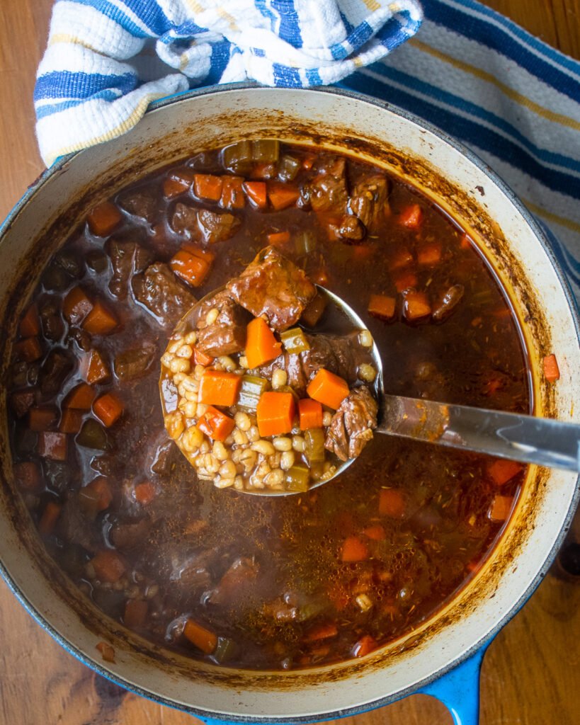 A ladle lifting beef barley soup out of a cast iron Dutch oven with a blue and yellow striped towel near by.