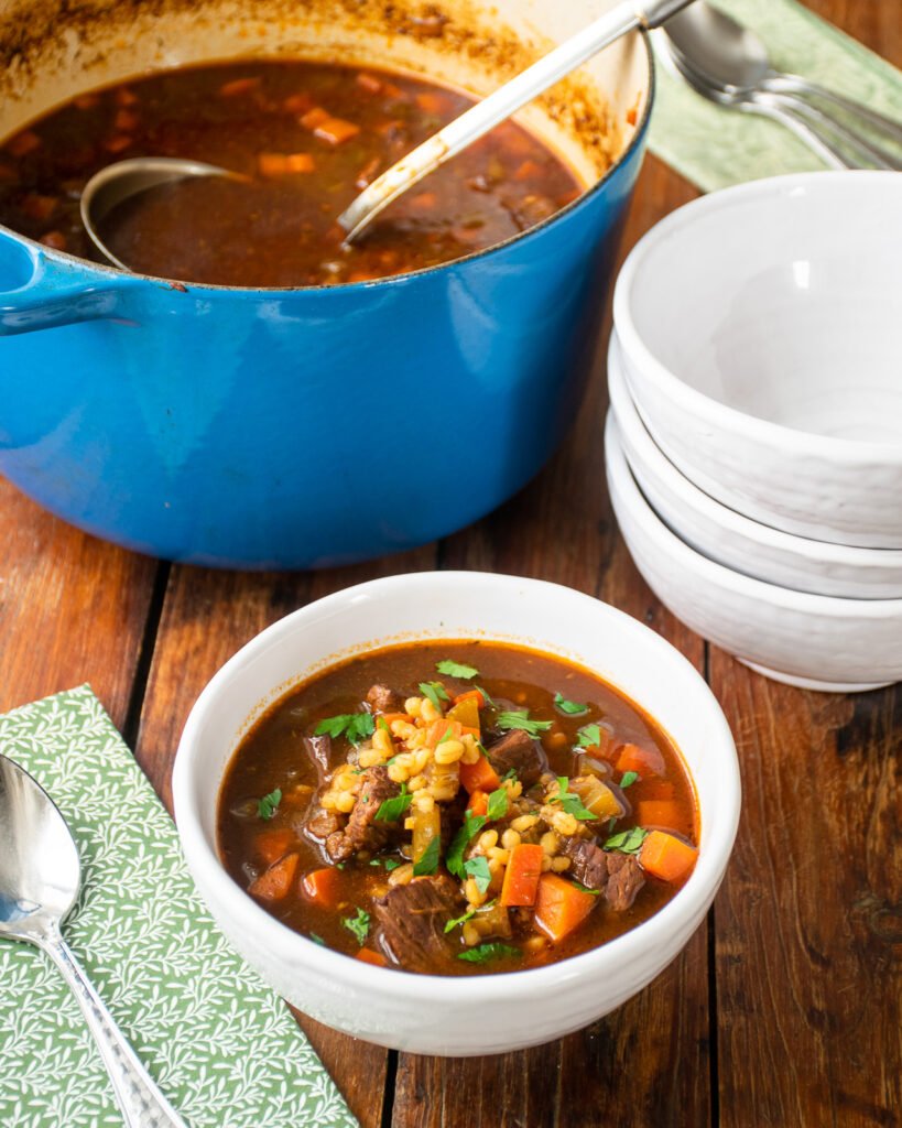A white bowl of beef barley soup in front of a stack of bowls and a blue cast iron Dutch oven.