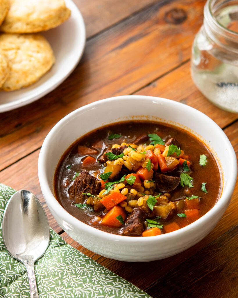 A white bowl of beef barley soup on a wooden table with green napkin, spoon and plate of biscuits.