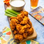 Air Fried Buffalo Cauliflower on a wooden board with carrot and celery sticks and blue cheese dressing, all on a blue countertop with a beer in the background.