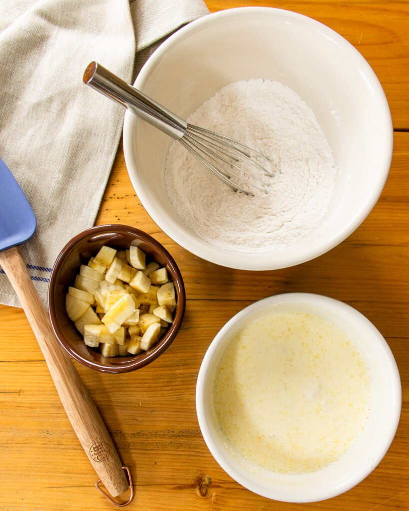 Three bowls on a table - a small bowl of chopped banana, a larger bowl with dry ingredients, a smaller bowl with wet ingredients - and a towel and blue spatula.