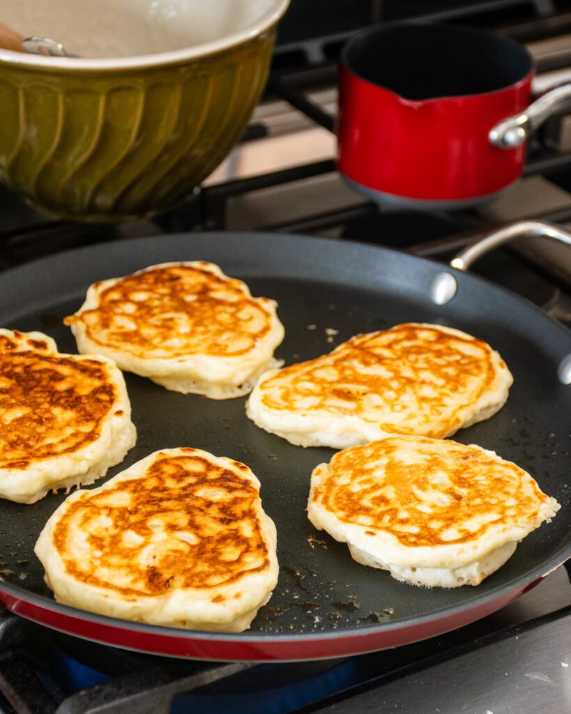 Pancakes cooking on a red griddle with a small red pot and a green bowl in the background.