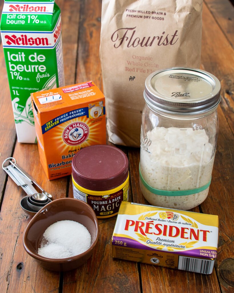 Ingredients on a table - buttermilk, baking soda, baking powder, salt, sugar, butter, sourdough starter and flour.