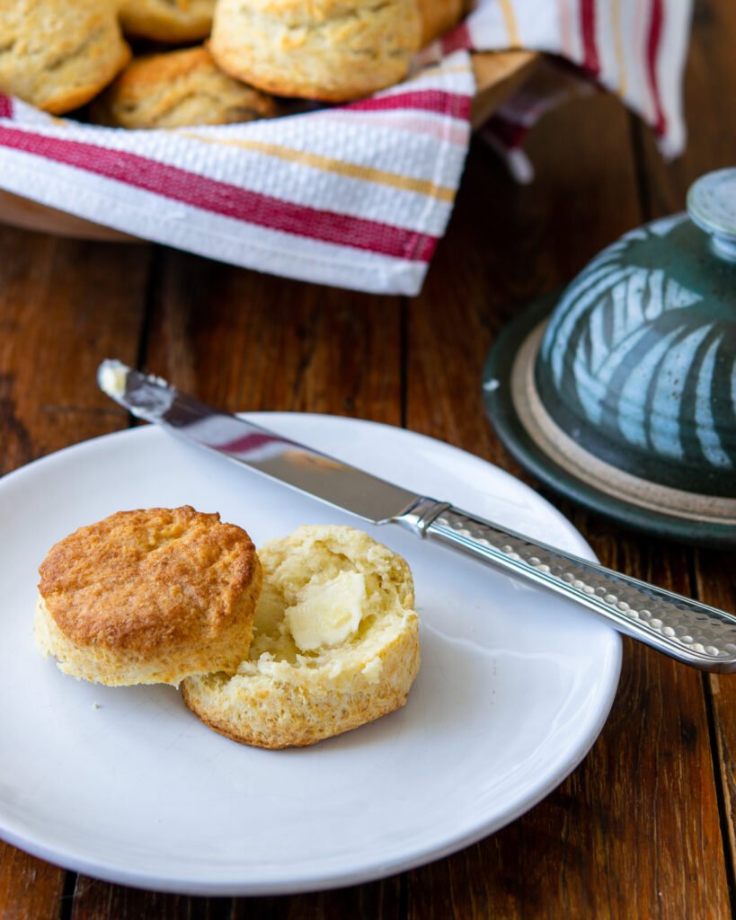 A sourdough buttermilk biscuit on a white plate with butter knife and more biscuits in the background.