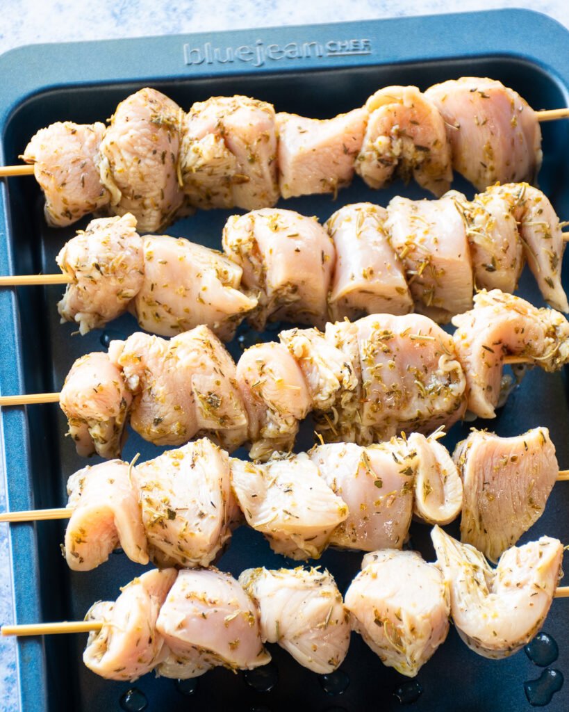 Raw marinated chicken on skewers in a blue baking pan.