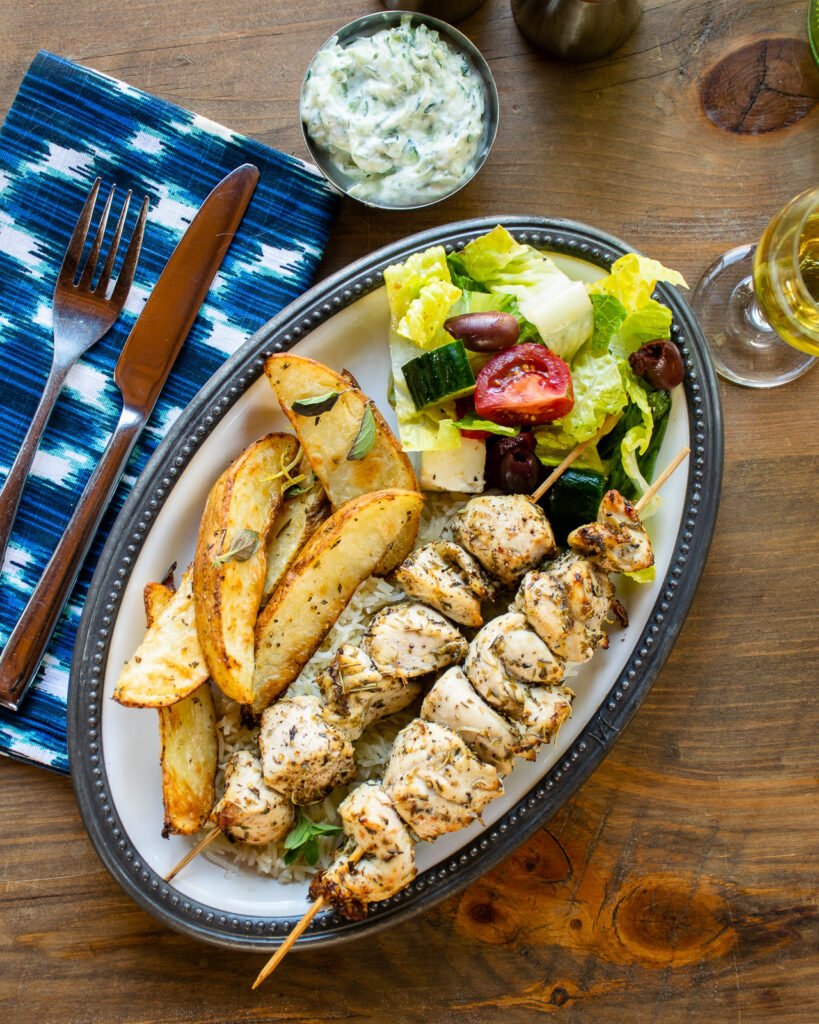Chicken souvlaki on a plate with Greek potatoes and salad with a place setting on a wooden table.