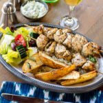 Chicken souvlaki on a plate with Greek potatoes and salad with a place setting on a wooden table.