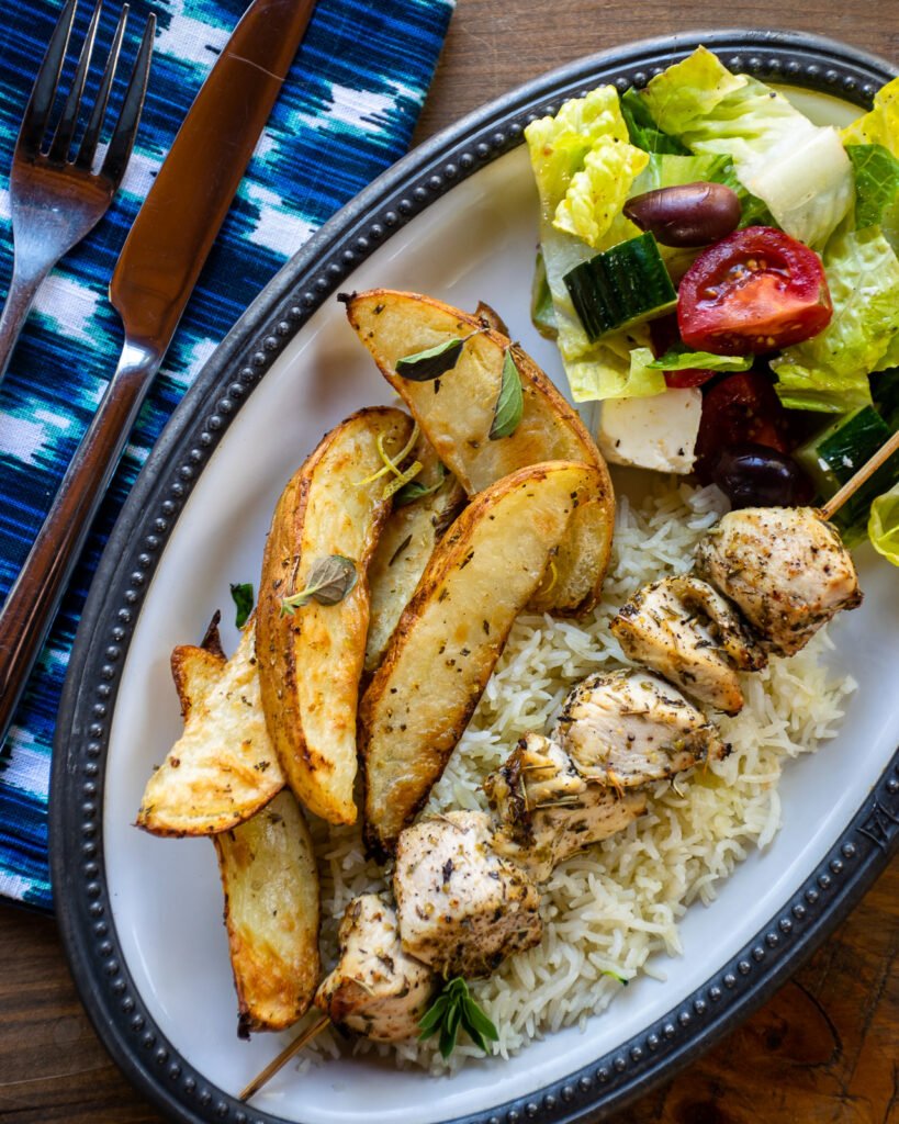 Greek potato wedges on an oval plate with chicken souvlaki and Greek salad.
