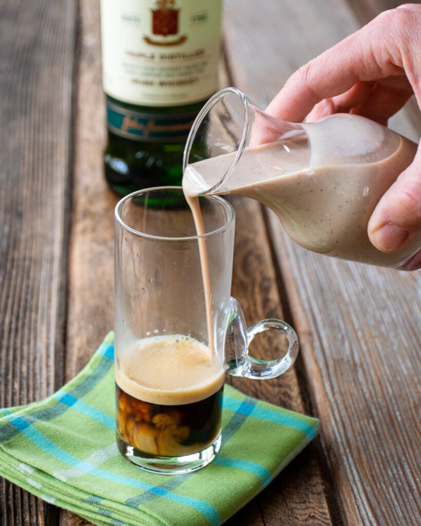 A hand pouring homemade Irish cream from a small carafe into a glass with coffee on a green plaid napkin.