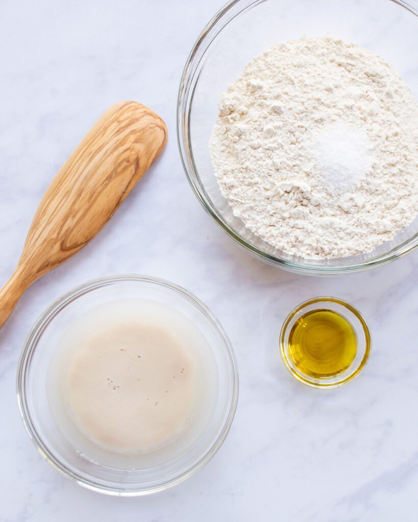 Yeast proofing in a glass bowl with a small bowl of olive oil and a large bowl of flour and salt, all next to a wooden spoon on a marble countertop.