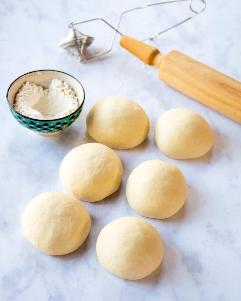 Six pita bread dough balls on a counter with a flour wand, a bowl of flour and a rolling pin.
