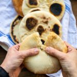 Hands breaking a homemade pita bread in half over a plate of finished pita breads.