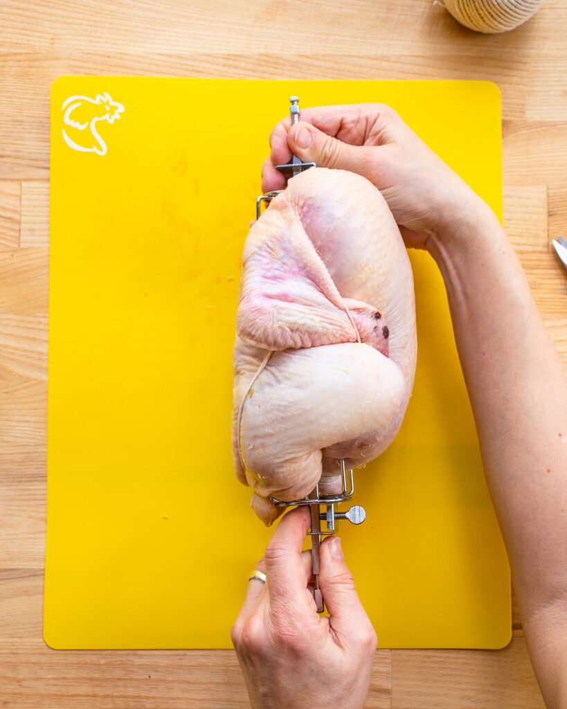 Two hands holding a trussed chicken on a rotisserie spit above a yellow cutting board.