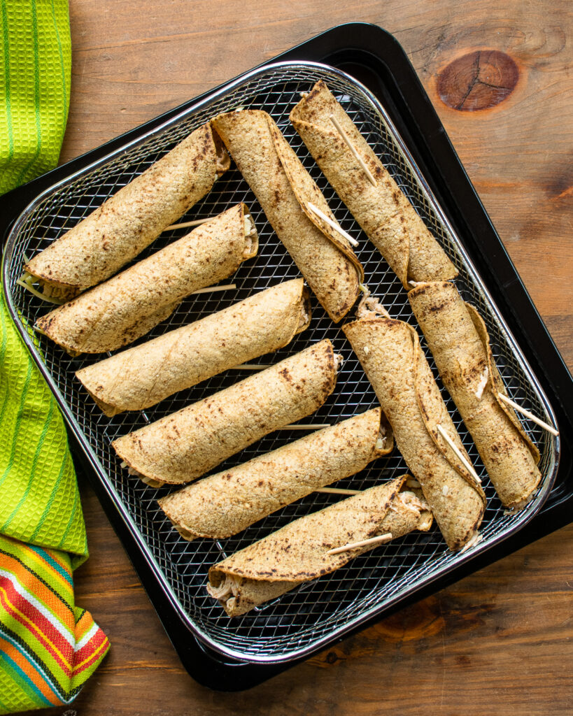 Tortillas rolled around a filling and closed with a toothpick sitting on an air fryer basket.