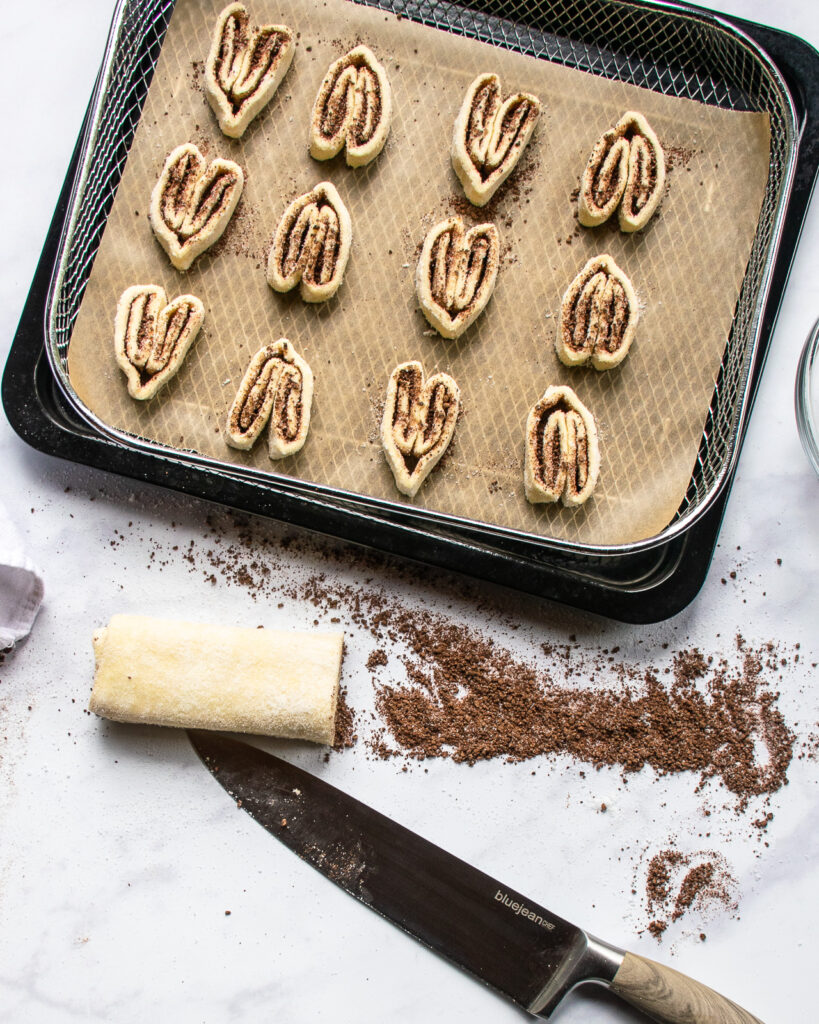 Chocolate palmiers on parchment paper on a baking tray with a log of puff pastry in front of it with a chef's knife.