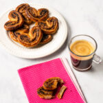 Chocolate Palmiers on a white plate with one palmier on a pink beverage napkin with an espresso.