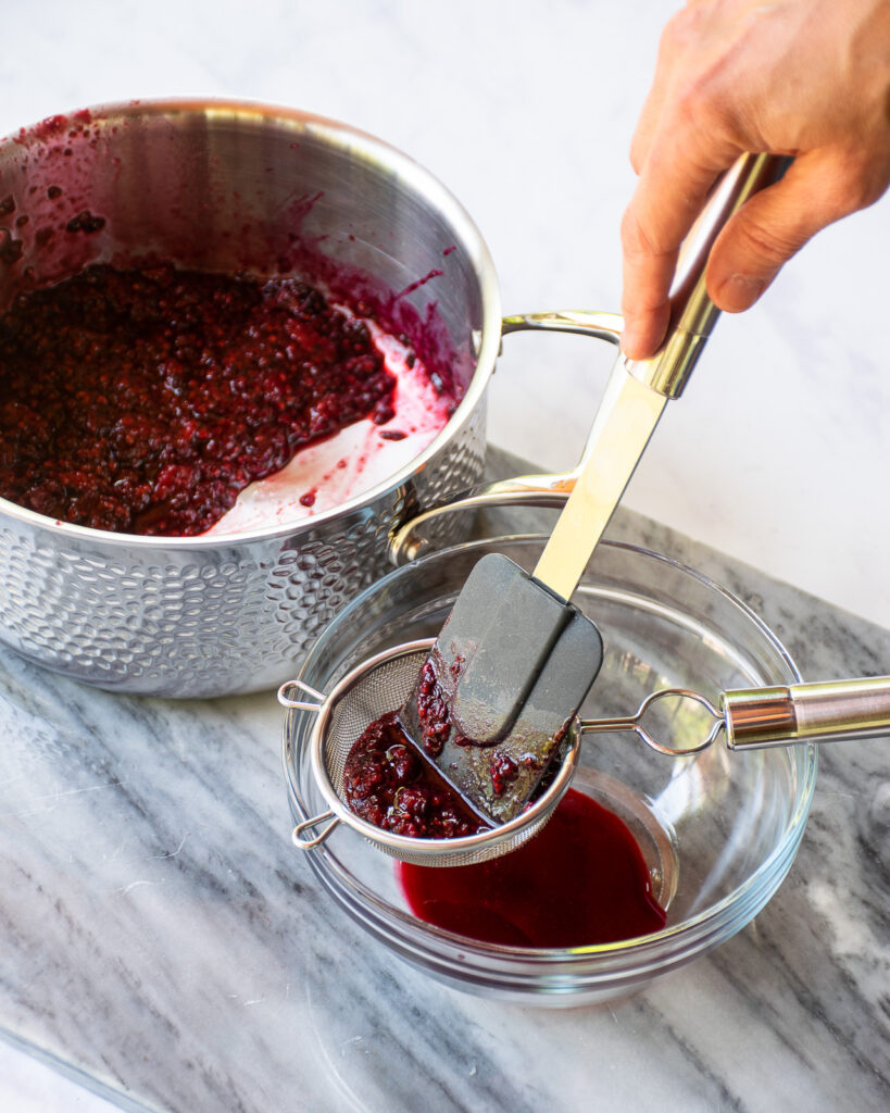 A hand pushing a blackberry purée through a fine strainer into a bowl with a saucepan in the background.