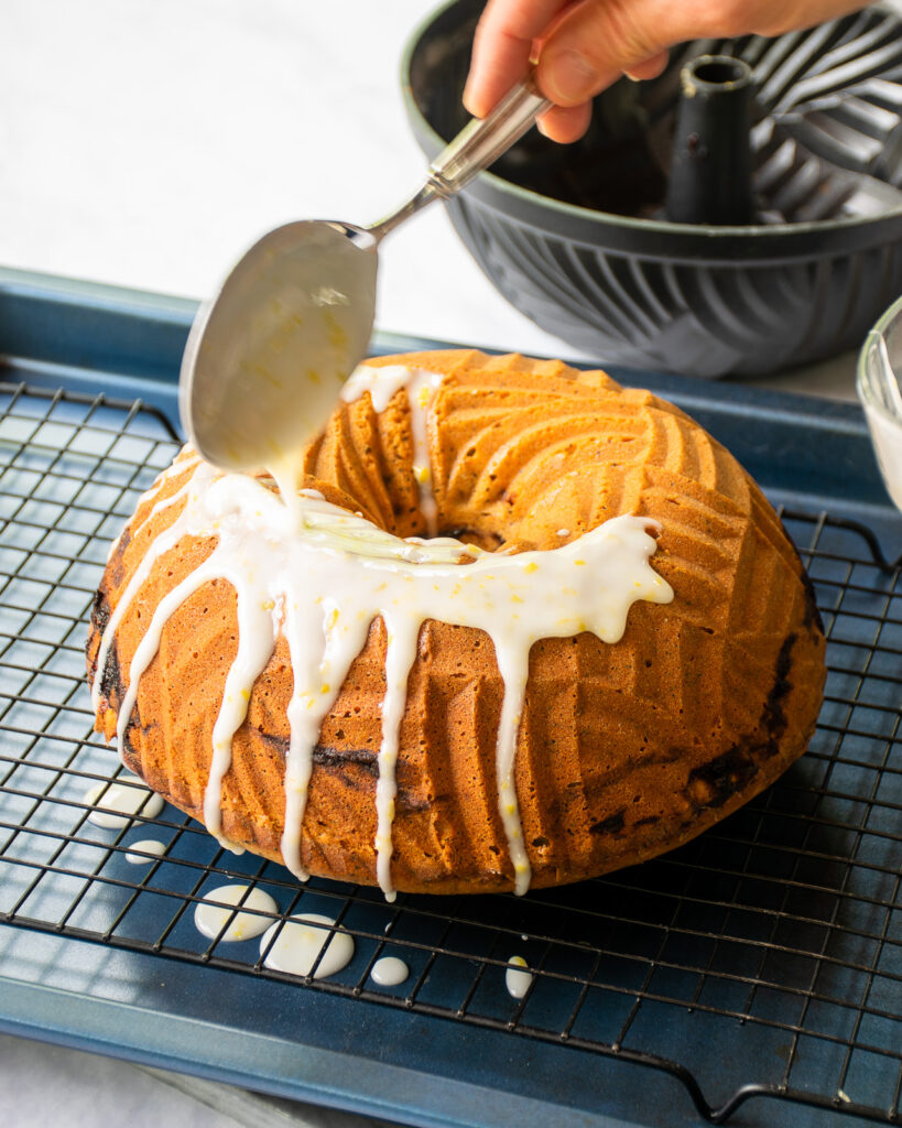 Spooning lemon glaze on top of lemon blackberry poppy seed bundt cake sitting on a cooling rack over a sheet pan.