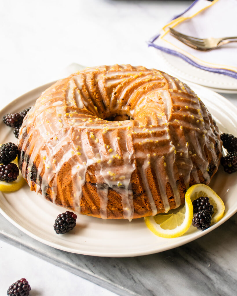 Lemon blackberry poppy seed Bundt® cake on a white plate with blackberries and lemon slices around the side.