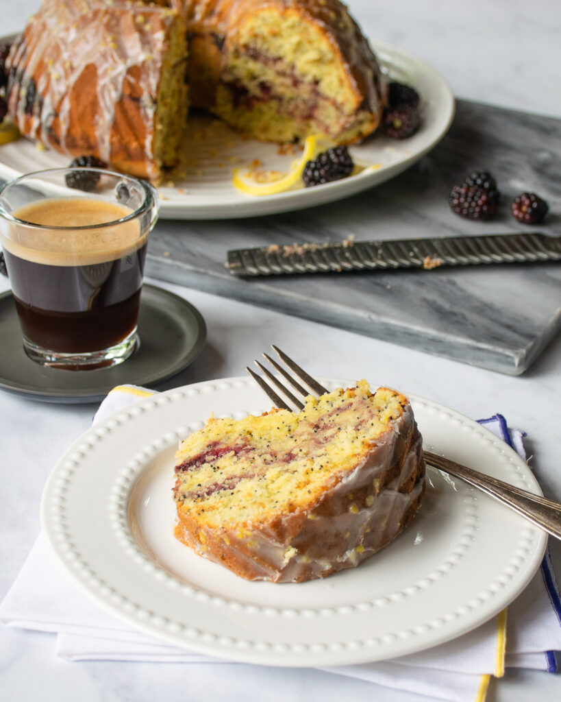 A piece of lemon blackberry poppy seed bundt cake on a white plate with coffee and the rest of the cake behind it.